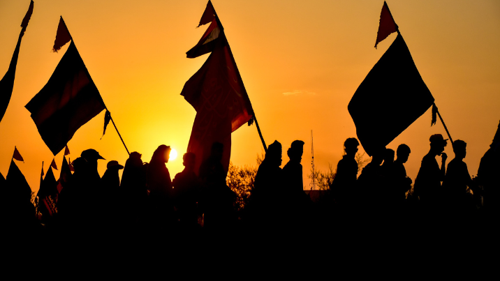 A photograph of the silhouette of pilgrims at dusk making their way to Karbala from Najaf. The sky glows warm yellow behind them, they carry flags and supplies for their journey.
